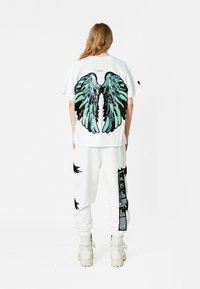 Camiseta blanca de gran tamaño con gráfico de alas verdes y negras en la parte posterior, combinada con pantalones blancos ajustados con acentos negros y bolsillos.