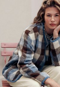 Kurtka z sztucznego futra w kratę w kolorach brązowym, niebieskim i kremowym, z niebieskim wykończeniem. Modelka pozuje na różowym fotelu.