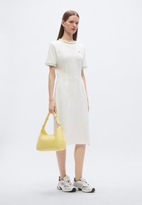 Λευκό φόρεμα μέχρι το γόνατο με κοντά μανίκια και πράσινη διακόσμηση, συνδυασμένο με κίτρινη τσάντα ώμου και λευκά αθλητικά παπούτσια με μαύρες λεπτομέρειες.