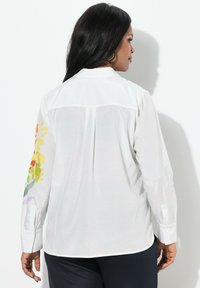 Camicia bianca a maniche lunghe con un motivo floreale su una manica, caratterizzata da un polsino con bottoni e un tessuto morbido e leggero.