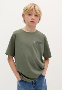 Olijfgroene T-shirt met korte mouwen van katoen, met een kleine grafische afbeelding op de borst, gecombineerd met blauwe denim jeans.