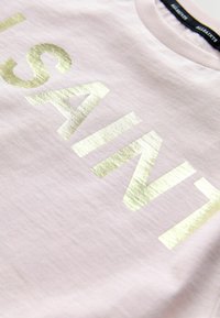 T-shirt en coton rose clair avec un grand texte en dorure indiquant "SAINT." Manches courtes et col rond. Texture lisse.