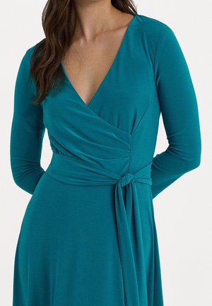 Kobieta w turkusowej, długorękawowej sukience kopertowej z dekoltem w serek i wiązaniem w talii, widziana od ramion do połowy uda na jednolitym tle.