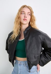 Μαύρο μπουφάν από συνθετικό δέρμα με φερμουάρ στο μπροστινό μέρος και ριμπέ κολάρο, φορεμένο πάνω από ένα πράσινο κοντό τοπ και μπλε τζιν σορτσάκια. Ορατή λεπτομέρεια λογότυπου.