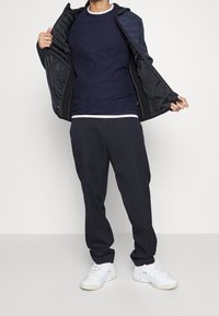 Mörkblå zip-up jacka med kviltade axlar, lager över en figurnära mörkblå skjorta. Matchas med lösa mörka byxor och vita sneakers.