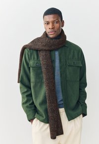 Groene wollen jas met knoopzakken, over een blauwe trui gedragen, gecombineerd met een bruine, gestructureerde sjaal. Lichtgekleurde broek maakt de look compleet.