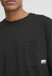 Black cotton t-shirt featuring a crew neck, short sleeves, and a left chest pocket with a white label displaying "11 PROJECT." Soft texture.