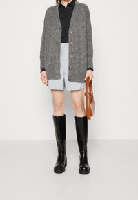 Grijze gebreide cardigan met houten knopen, zwart shirt, lichtgrijze op maat gemaakte shorts, zwarte kniehoge laarzen en een bruine handtas.