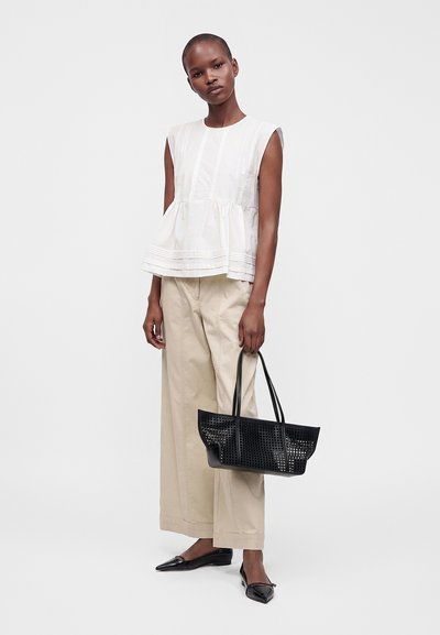 Femme portant un haut blanc sans manches et un pantalon beige à jambes larges, tenant un sac cabas noir tissé et portant des ballerines noires pointues.
