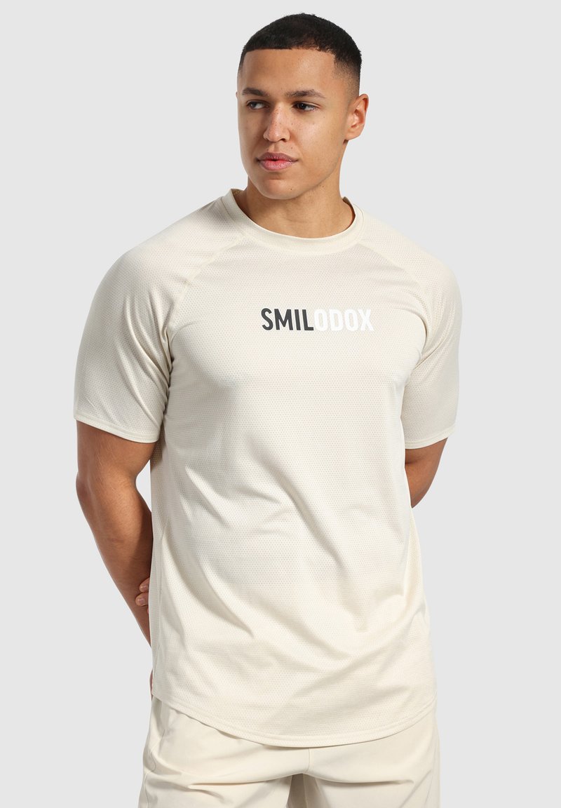 Camiseta deportiva beige con textura de malla transpirable, mangas cortas y el logo "SMILODOX" en blanco y negro centrado en el pecho.