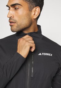 Black Terrex jacket with high collar, front zip, and branded logo. Smooth texture, fitted design highlighting the neck and shoulders.