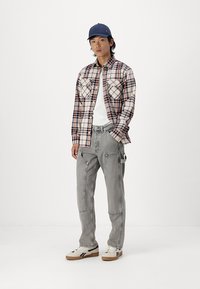 Geruite flanellen shirt in beige, marineblauw en rood met twee borstzakken, gecombineerd met grijze cargobroeken en witte sneakers met donkere accenten. Pet gedragen.