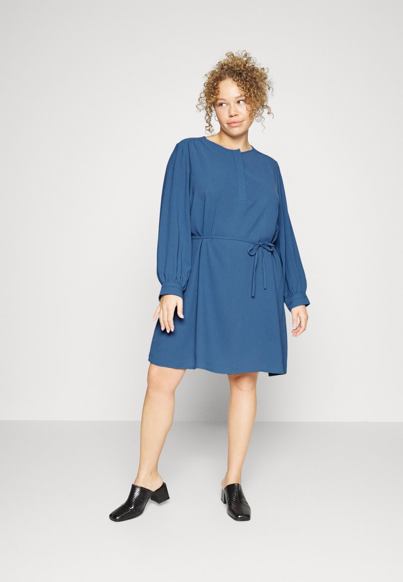 Vestido azul de manga curta com decote redondo, cintura elástica e detalhe de laço. Usado com sapatos pretos texturizados de deslizar. Tecido suave e corte descontraído.