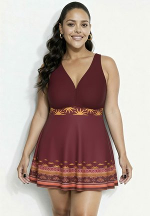 Donna sorridente con lunghi capelli scuri indossa un vestito senza maniche color bordeaux con scollo a V profondo e orlo decorativo a motivo giallo-arancione e fascia in vita.