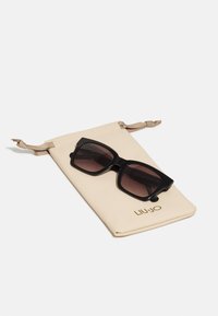 Occhiali da sole neri oversize con lenti sfumate su un sacchetto con cordoncino beige, dotato di un logo dorato. Texture liscia, forma rettangolare.