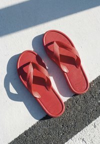Paire de tongs rouges Havaianas reposant sur une surface asphaltée blanche et noire texturée, avec la lumière du soleil projetant des ombres.