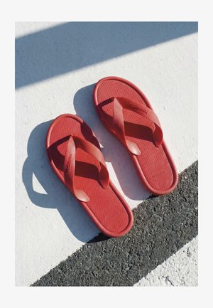 Ein Paar rote Havaianas Flip-Flops, die auf einer weiß-schwarz strukturierten Asphaltoberfläche liegen, mit Sonnenlicht, das Schatten wirft.