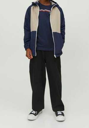 Granatowa i beżowa kurtka zapinana na zamek z kapturem, zestawiona z granatowym T-shirtem z poziomą czerwoną paskiem oraz luźnymi czarnymi spodniami i trampkami.