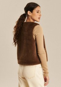 Kobieta z długimi brązowymi włosami związanymi w kucyk, ubrana w bezrękawnik z brązowego futra, beżową bluzkę z długim rękawem i kremowe spodnie, stojąca bokiem.