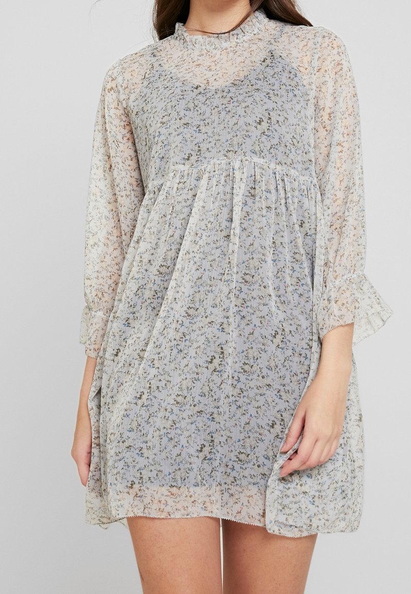 Robe fleurie transparente à coupe ample, de couleur bleu clair et crème, taille froncée, manches à volants et motifs floraux délicats sur toute la surface.