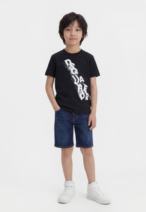 Νέος αγόρι στέκεται με τα χέρια στις τσέπες, φορώντας μαύρο γραφικό μπλουζάκι, μπλε τζιν σορτς και λευκά αθλητικά υψηλού πάτου.