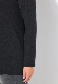 Langärmliges Shirt aus dunkelgrauem Stoff. Weiche Textur, abgerundete Ärmelbündchen, leicht tailliert in der Taille. Haut ist am Handgelenk sichtbar.
