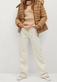 Brun pufferjakke med hætte og ribbet beige sweater, parret med hvide vide bukser og hvide sneakers med grønne detaljer.
