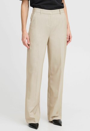 Beige gerade geschnittene Hosen mit Bügelfalte vorne, Seitentaschen, Gürtelschlaufen, kombiniert mit schwarzen spitzen Schuhen und schwarzem Oberteil vor einfachem Hintergrund.