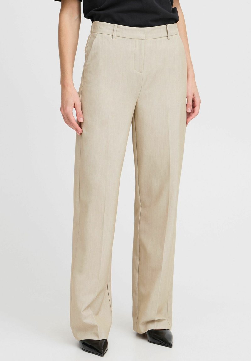 Pantalon beige à jambe droite avec pli sur le devant, poches latérales, passants pour ceinture, porté avec des chaussures noires pointues et un haut noir sur fond uni.