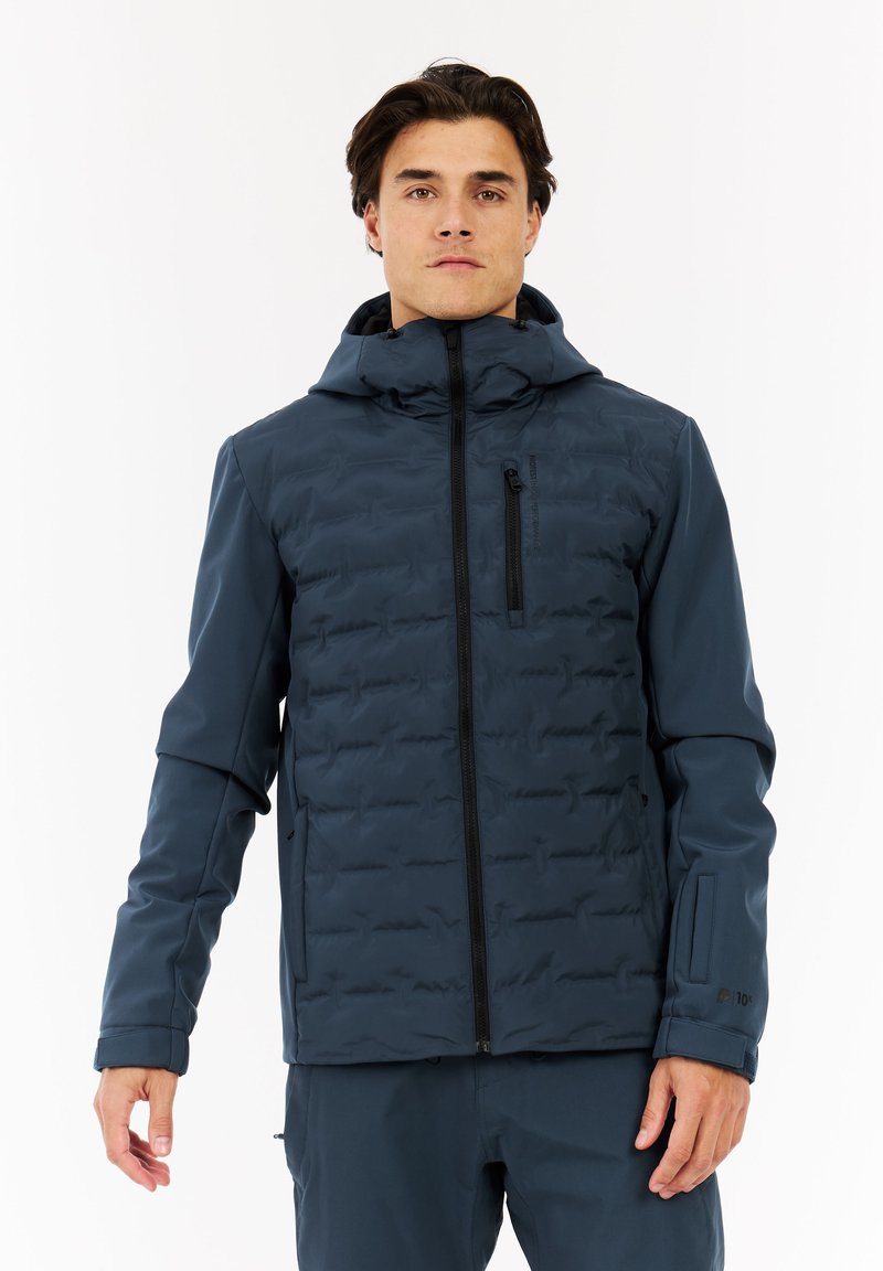 Man in een donkerblauwe geïsoleerde jas met zwarte rits en capuchon, staande tegen een witte achtergrond.