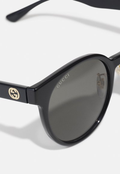 Czarne okrągłe okulary przeciwsłoneczne z ciemnymi szkłami, złote logo Gucci na zausznicy oraz "GUCCI" wyryte na krawędzi szkła.