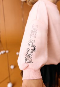 Ljusrosa sweatshirt med texten "TRICK OR TREAT" på ärmen och en spindelillustration. Har ribbade muddar och en avslappnad passform.