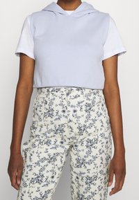 Haut sans manches bleu clair avec capuche porté sur un t-shirt blanc, associé à un pantalon taille haute avec un motif floral bleu sur fond crème.