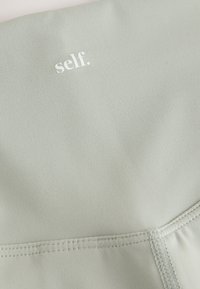 Heledast rohelised jooga püksid, millel on sile tekstuur. Vöökohal valge logoga „self.” ja õmbluste ääres tugevdatud õmblused.