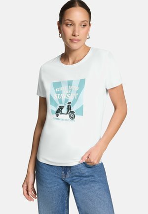 Femme portant un t-shirt blanc avec un graphisme d'un scooter et des rayons de soleil ainsi que le texte "Partez vers le coucher du soleil, escapade d'été," assorti à un jean bleu.