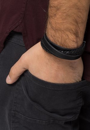 Męska ręka z czarną plecioną skórzaną bransoletką spoczywająca w kieszeni czarnych spodni, ubrana w ciemnoczerwoną koszulę.