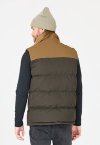 Padded vest in olive green and tan, featuring a high collar, sleeveless design, and a matte finish. Paired with a light beige beanie.
