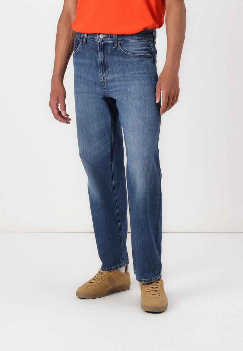 Jeans en denim bleu avec une coupe droite, un design à cinq poches et des détails de couture visibles. Associés à des baskets beiges et une chemise orange.