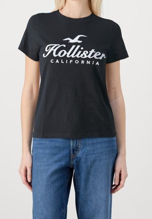 Femme portant un t-shirt noir Hollister California et un jean bleu, debout contre un fond clair uni.