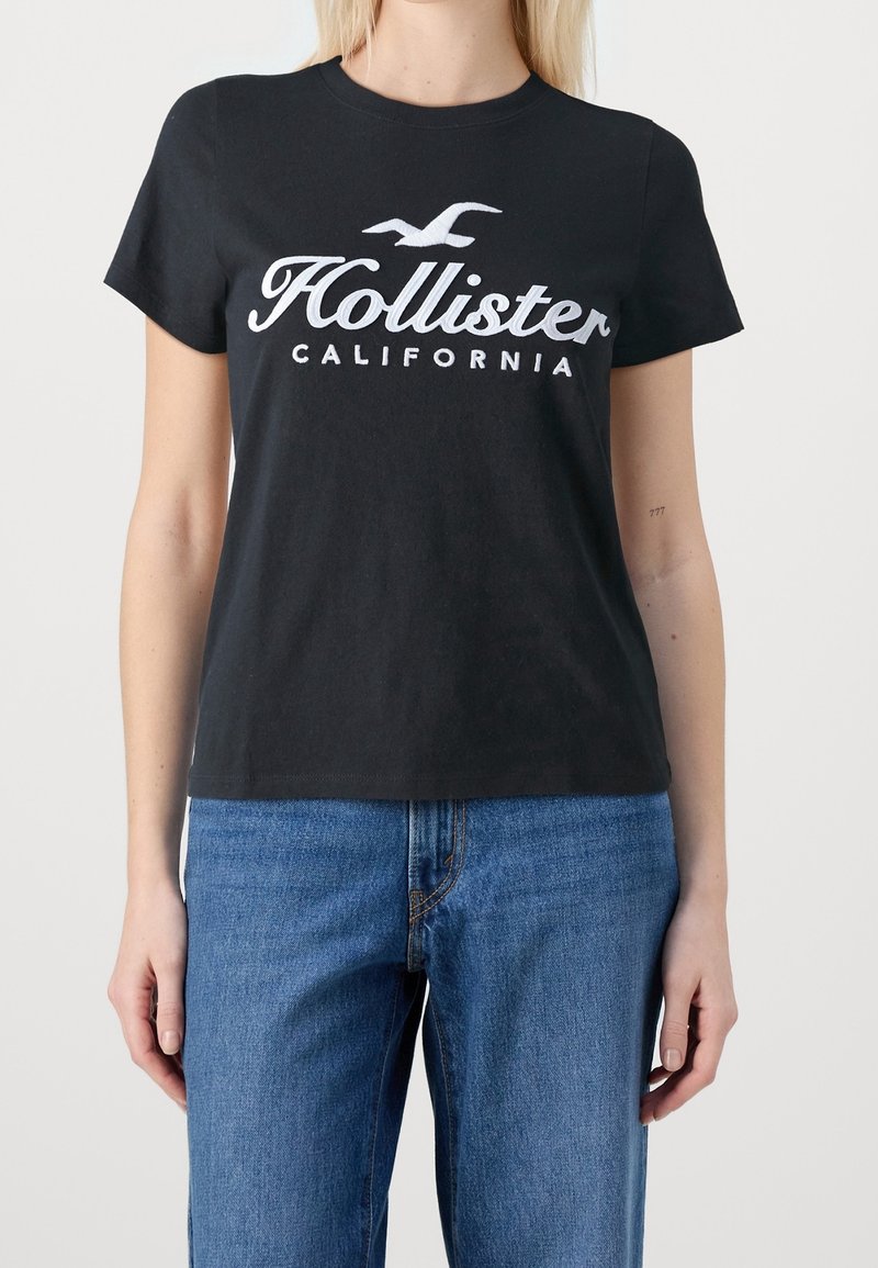 Kobieta ubrana w czarną koszulkę Hollister California i niebieskie dżinsy, stojąca na jasnym, jednolitym tle.