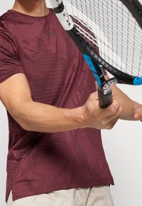 Mörk röd idrottströja med korta ärmar, med ett mönstrat textur. Bäraren håller en tennisracket och visar en sportig design.