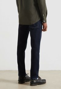 Jeans di denim blu scuro con taglio dritto, abbinati a una camicia verde e scarpe nere, visti da dietro.