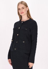 Cardigan in maglia a treccia navy con scollatura rotonda, due tasche frontali e bottoni in tonalità oro. Presenta polsini e orlo a coste per un tocco di texture in più.