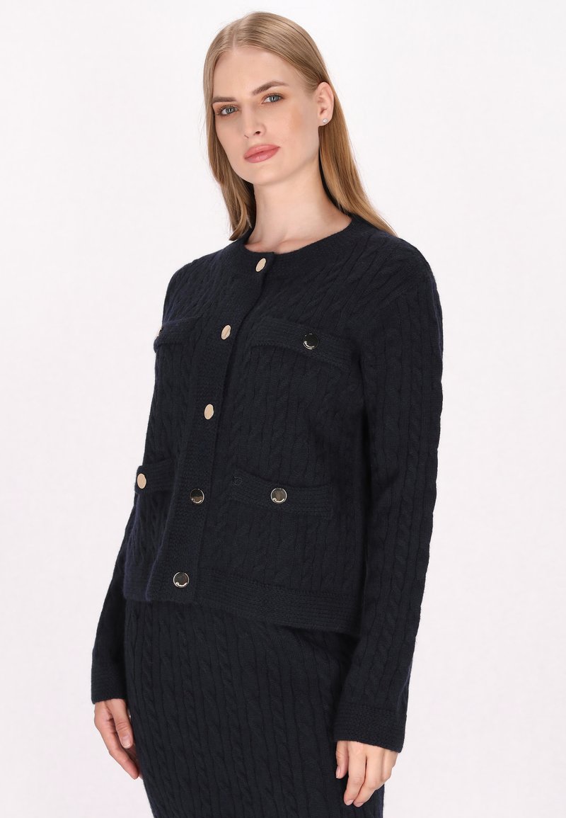 Cardigan in maglia a treccia navy con scollatura rotonda, due tasche frontali e bottoni in tonalità oro. Presenta polsini e orlo a coste per un tocco di texture in più.