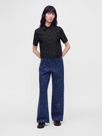 Femme aux longs cheveux noirs portant une chemise noire à manches courtes boutonnée et un jean bleu à jambes larges avec de petits motifs blancs, debout les mains derrière le dos.