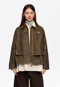 Zielona kurtka w kratę z zamkiem z przodu, z brązowym kołnierzem z kordurowego materiału i kontrastowym przeszyciem. Model nosi pod nią jasny golf.