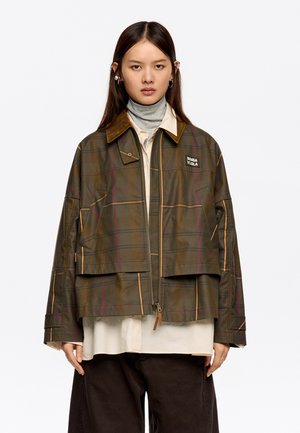 Chaqueta verde a cuadros con cremallera frontal, que presenta un cuello de pana marrón y costuras en contraste. El modelo lleva una camiseta de cuello alto clara debajo.