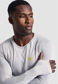 Maglietta sportiva a maniche lunghe di colore grigio chiaro con motivo a trama, caratterizzata da un logo verde sul petto e dettagli con foro per il pollice nei polsini.