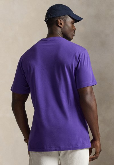 Mand iført en lilla t-shirt med korte ærmer og en mørkeblå kasket, stående med ryggen mod en neutral baggrund.