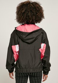 Zwarte en roze windjack met capuchon, voorzien van elastische manchetten en onderkant, plus witte accenten op de mouwen. Soepele textuur, ontspannen pasvorm.